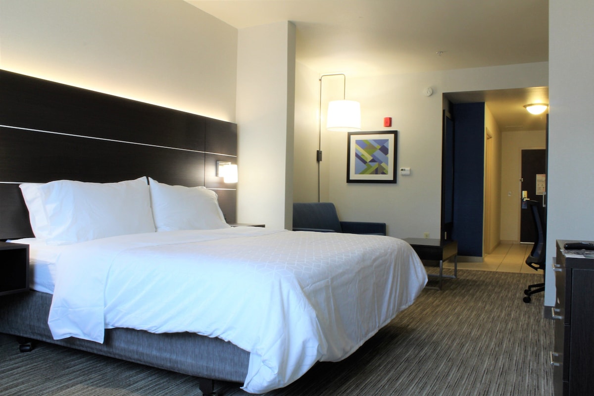 A hotel room with a bed, bedside table, lamp, chair, desk, and a door leading to another room.