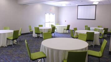 A conference room with multiple round tables and green chairs, carpeted floor, and a projector screen on the wall.