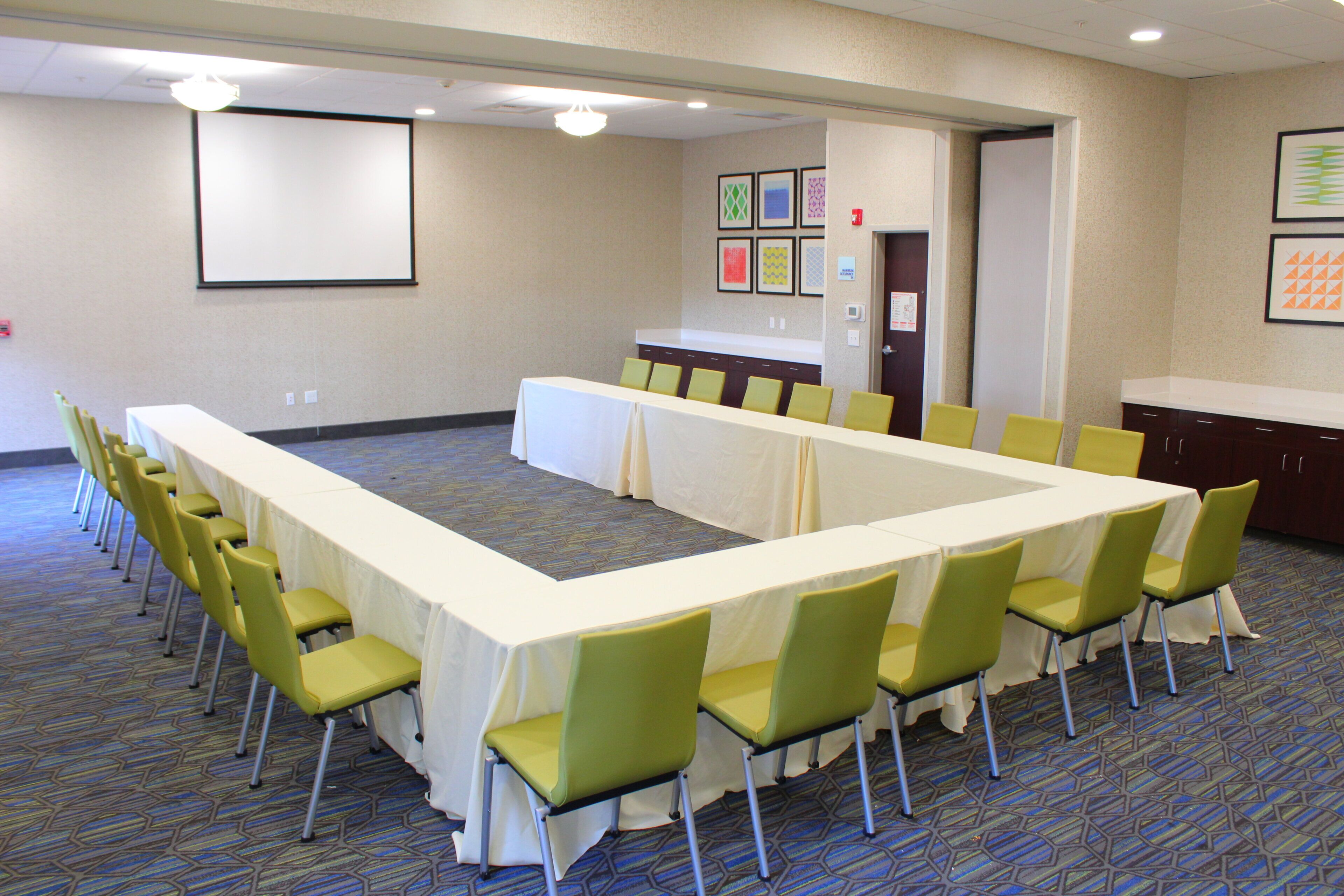 A conference room with a long table, chairs, a projector screen, and a kitchenette area.