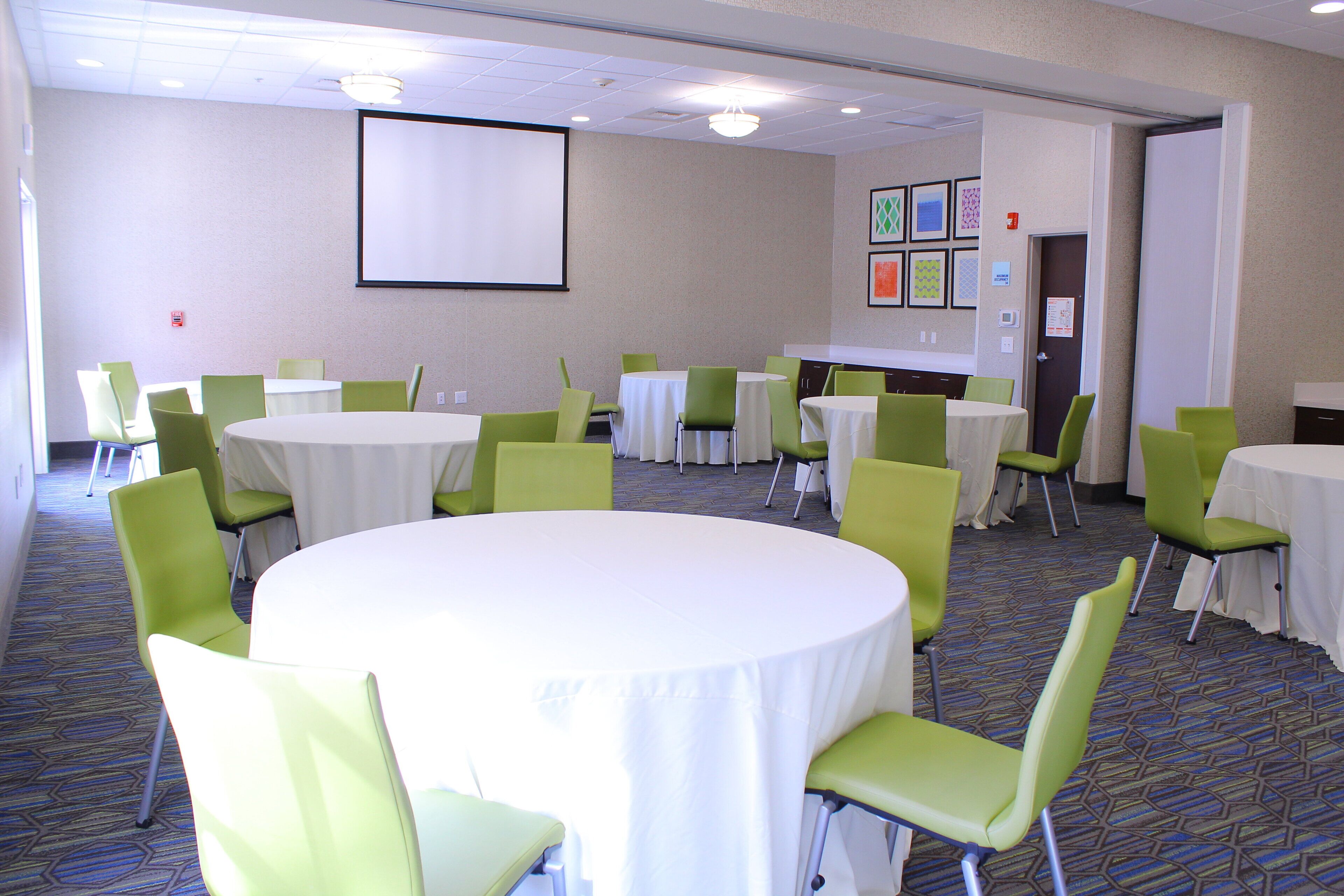 A conference room with multiple tables and chairs, a projector screen, and framed pictures on the wall.