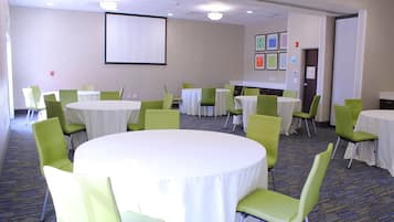 A conference room with multiple tables and chairs, a projector screen, and framed pictures on the wall.