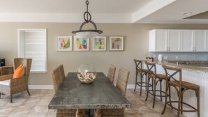A dining area with a dark table, wicker chairs, and a glass bowl on the table. There are white cabinets and a kitchen area in the background. - Hodnett Cooper St Simons Grand - 3 Bedroom (St. Simons Island)