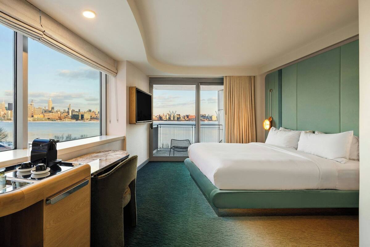 Habitación de hotel con una cama grande, un escritorio con cafetera, televisor y vistas a la ciudad a través de amplios ventanales.