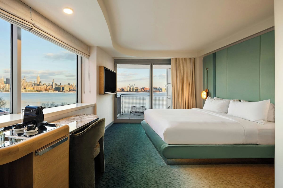 Une chambre d’hôtel avec un grand lit, un bureau avec une machine à café, une télévision et une vue sur le paysage urbain grâce à de grandes fenêtres.