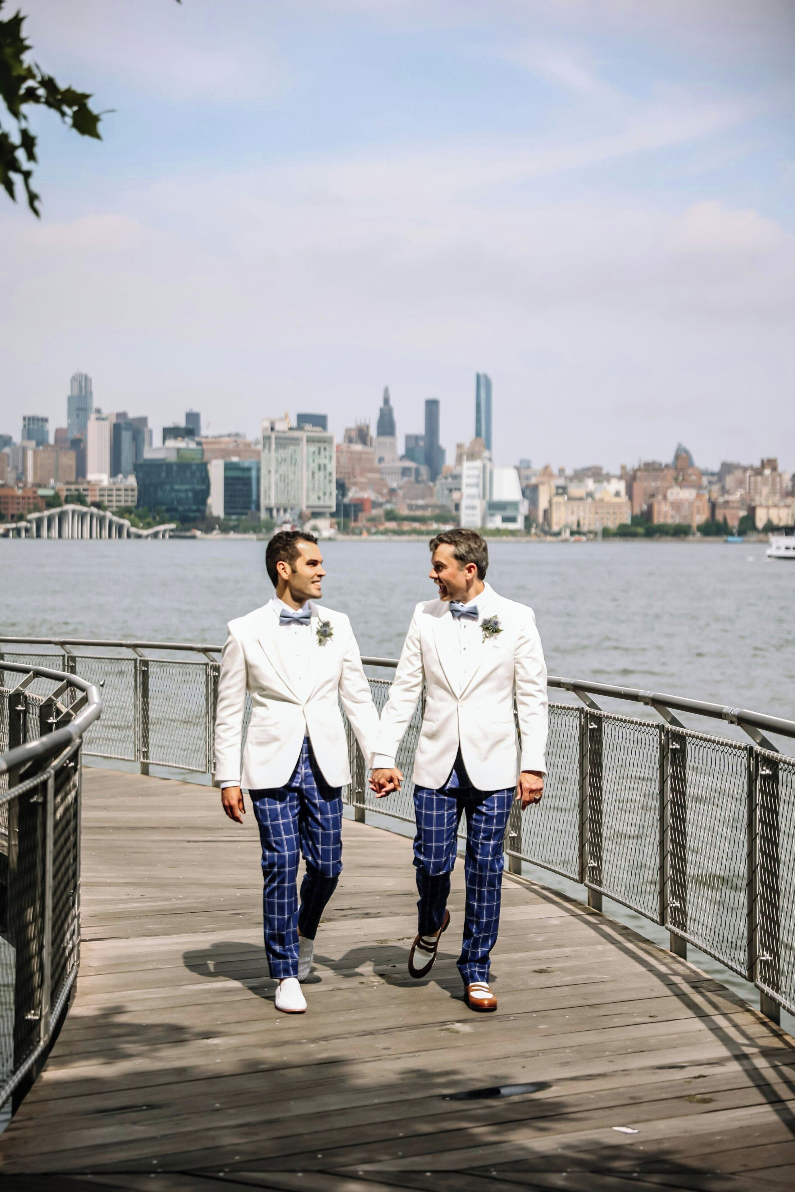 Dos hombres con trajes blancos caminando sobre una pasarela de madera con un horizonte urbano al fondo.