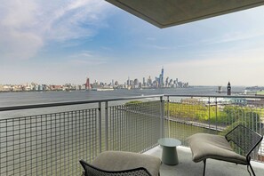 Un balcon offrant une vue sur les gratte-ciel d’une ville, avec une rivière en contrebas, agrémenté d’une chaise moderne et d’une petite table.