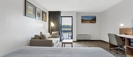 A hotel room with a bed, a sofa, a desk, and a chair. There is a window with a view of greenery and a door leading to an outdoor balcony.