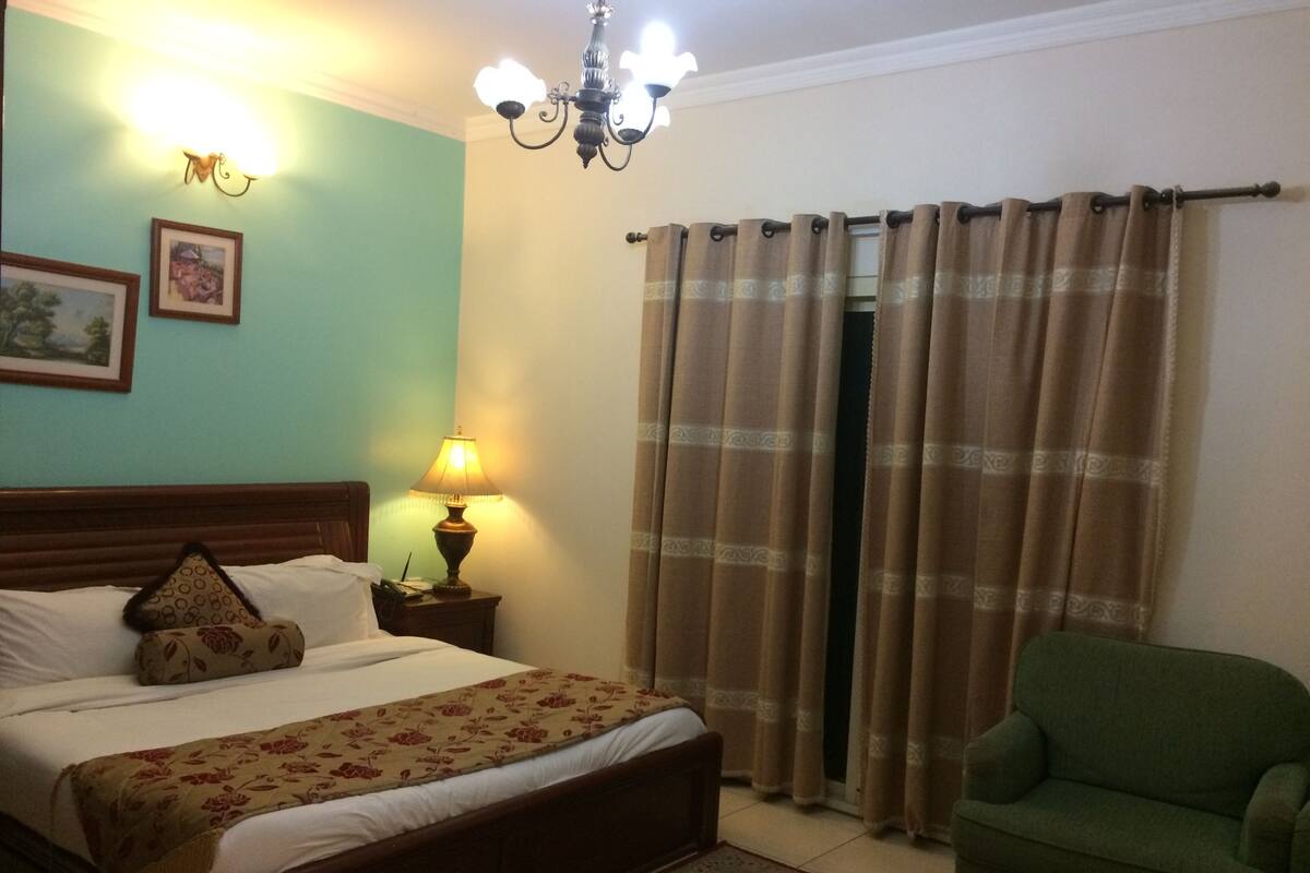 A bedroom with a bed, a nightstand, a lamp, a green armchair, and a window with curtains.