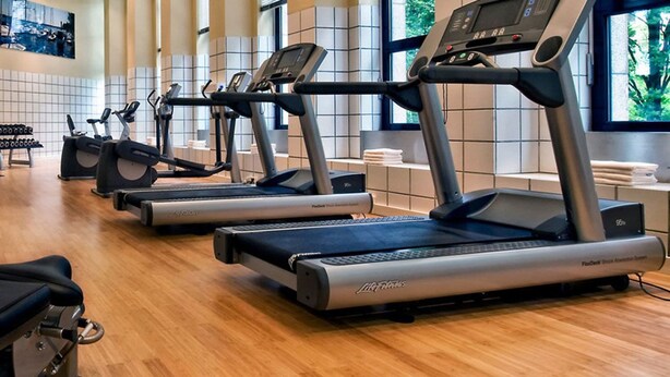 Varias caminadoras en un gimnasio con piso de madera y ventanas amplias.