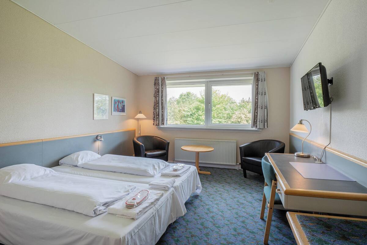 A hotel room with two beds, a desk, a chair, a small table, a TV, and a window with curtains.