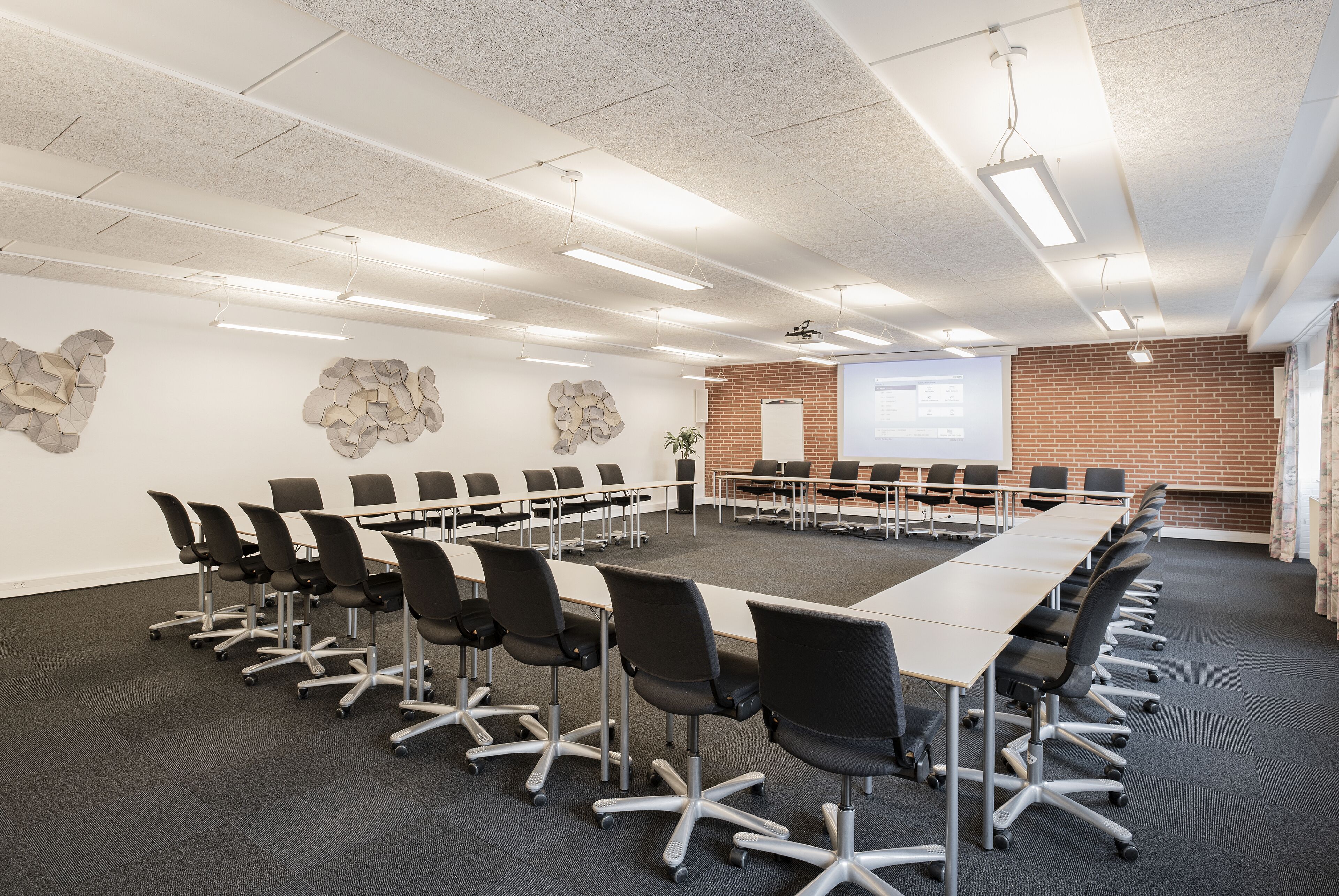 Un salón de conferencias con una mesa larga, sillas negras, una pantalla de proyector y una pared de ladrillo.
