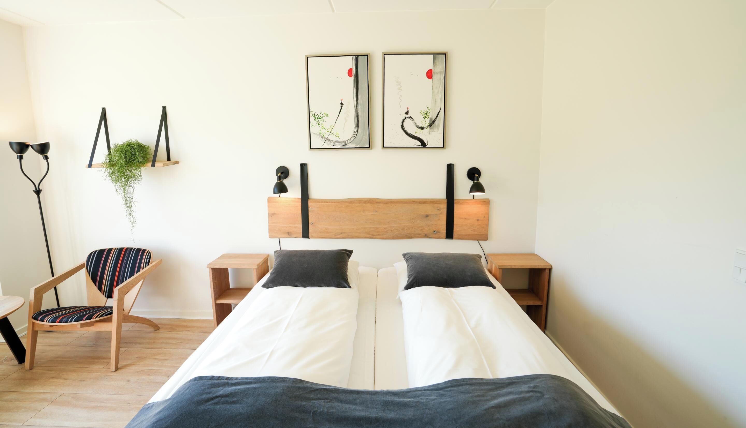 Un letto matrimoniale con testiera in legno, due comodini, una poltrona con cuscino a righe, una lampada da terra e due opere d'arte appese al muro.
