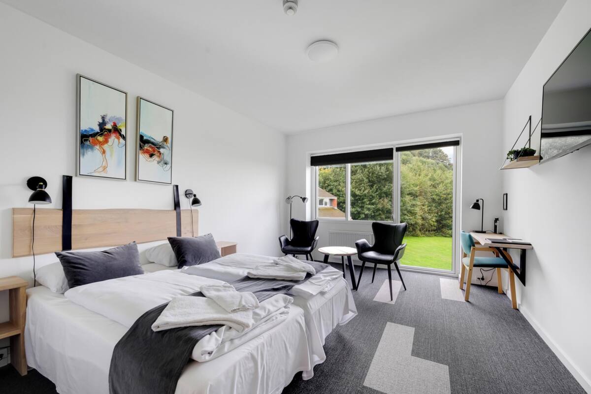 A modern hotel room with a large bed, two chairs, a small table, a desk, a TV, and artwork on the wall.