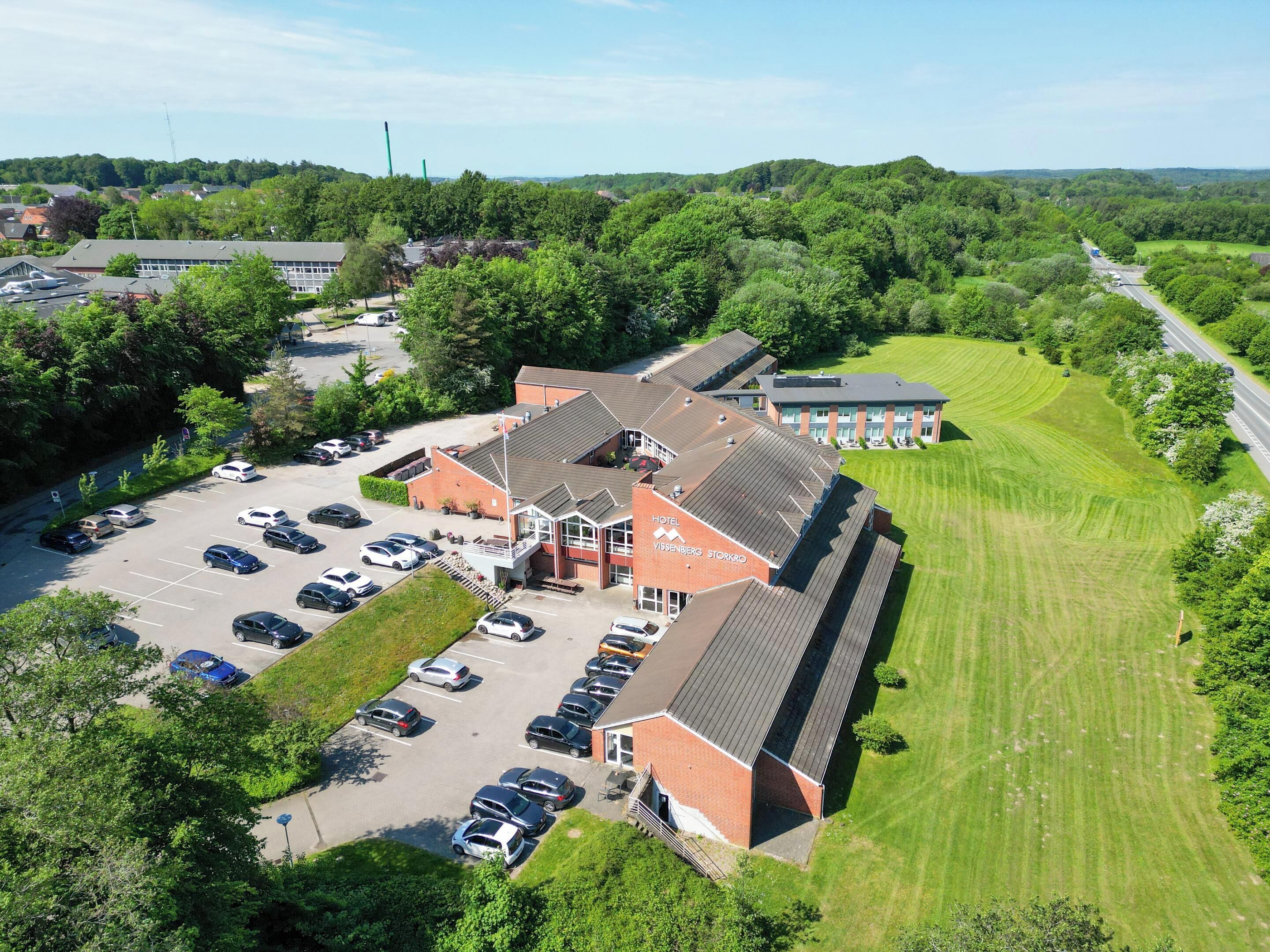 An aerial view of a hotel with a parking lot, surrounded by greenery and a road.