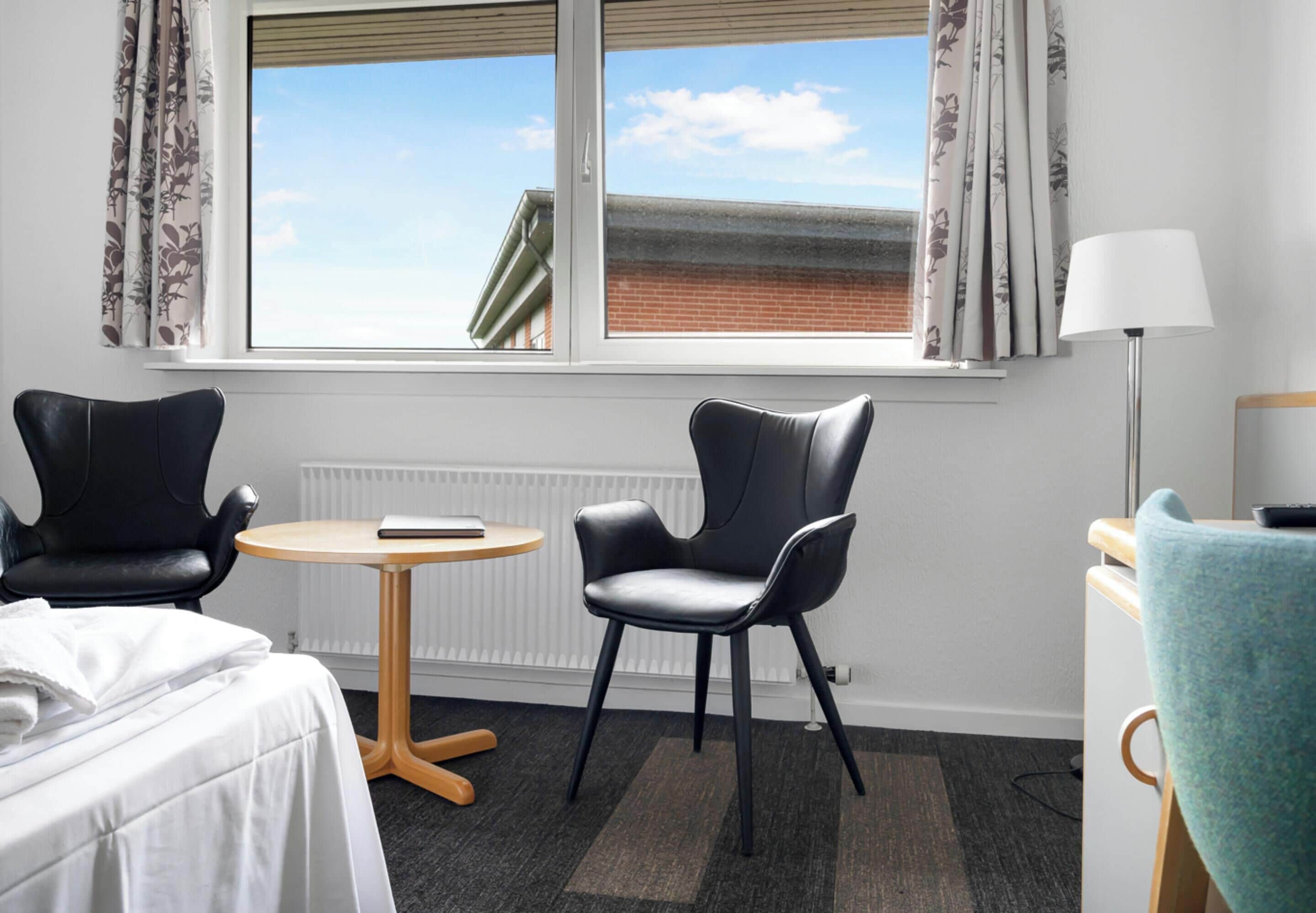 A hotel room with two black leather chairs, a small round table, a bed with white linens, a lamp, and a view of a building through a window with curtains.