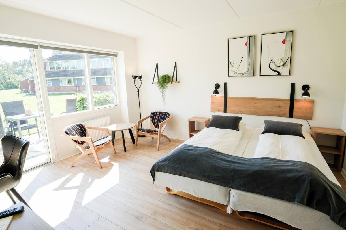 A modern bedroom with a large bed, a small table, two chairs, a window, and artwork on the wall.