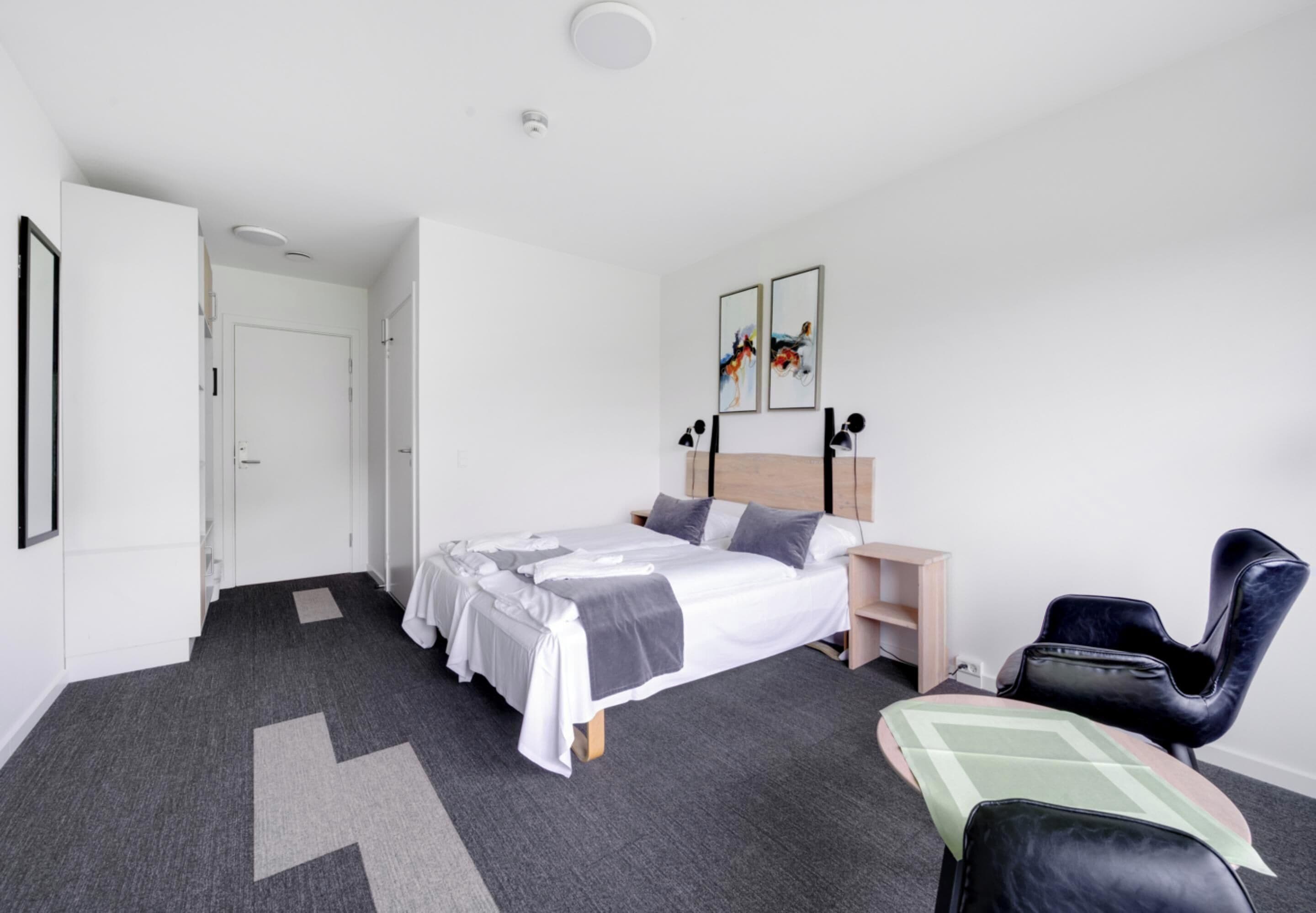 Quarto de hotel moderno com uma cama bem arrumada, uma escrivaninha, uma cadeira e uma porta que dá para outro quarto.