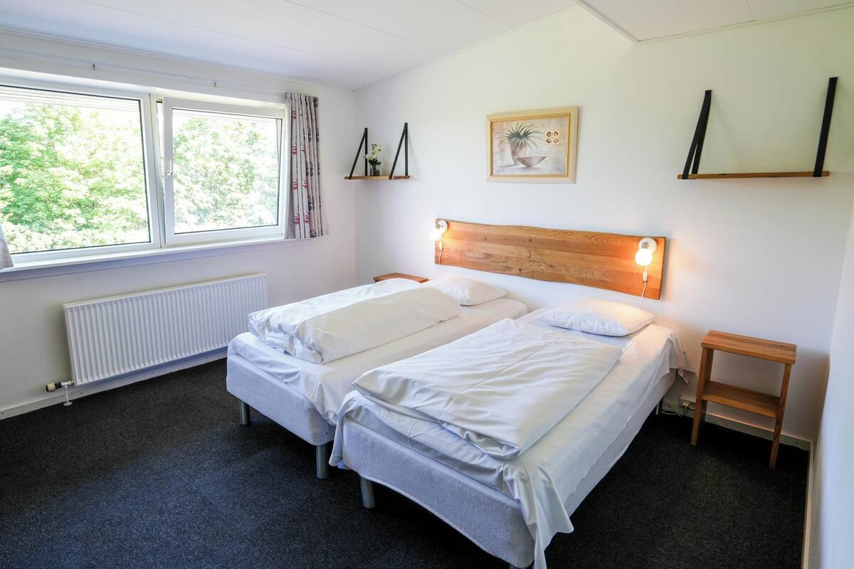 A double room with two beds, a radiator, a window with curtains, and a small bedside table.