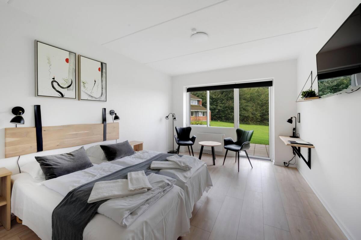 A modern hotel room with a large bed, two chairs, a small table, and a large window offering a view of the outdoors.