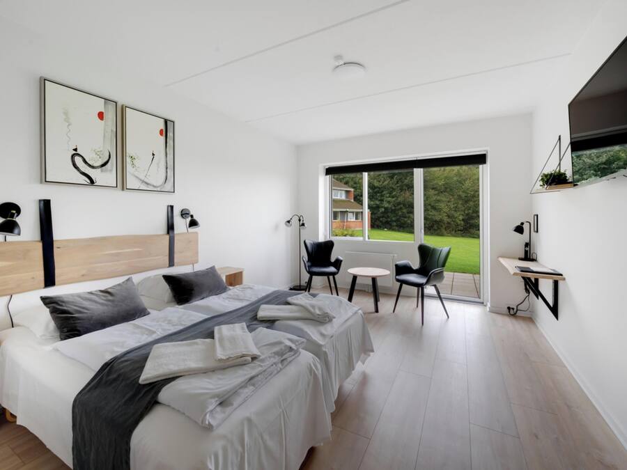 Une chambre d’hôtel moderne, dotée d’un grand lit, de deux fauteuils, d’une petite table et d’une grande fenêtre donnant sur l’extérieur.
