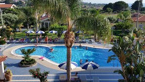 A swimming pool with lounge chairs and umbrellas, surrounded by palm trees and a view of houses in the background. - Hotel Damanse (Ricadi)
