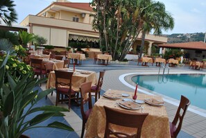 An outdoor dining area with tables and chairs, a pool, and a building with a tiled roof. - Hotel Damanse (Ricadi)