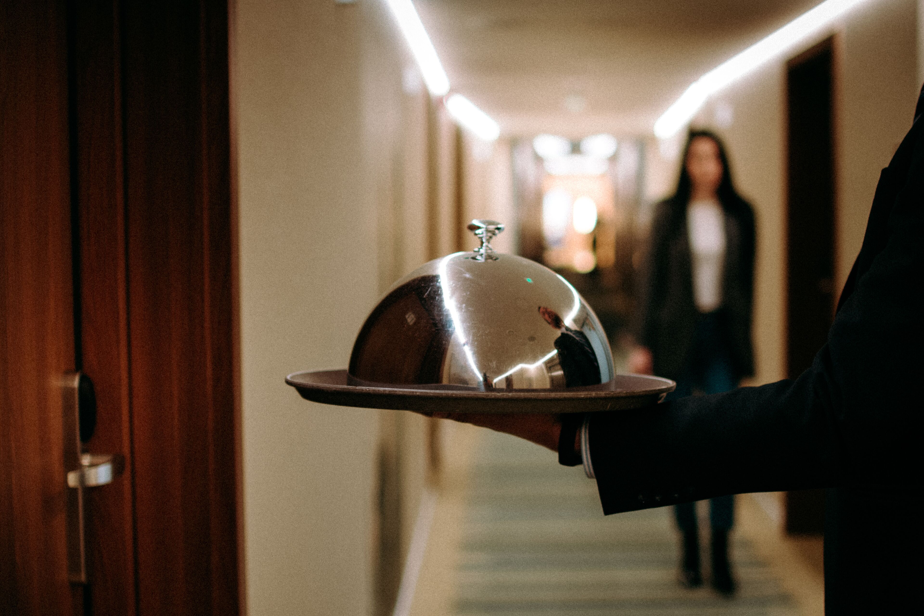 A person carrying a tray with a covered dish in a hotel hallway.