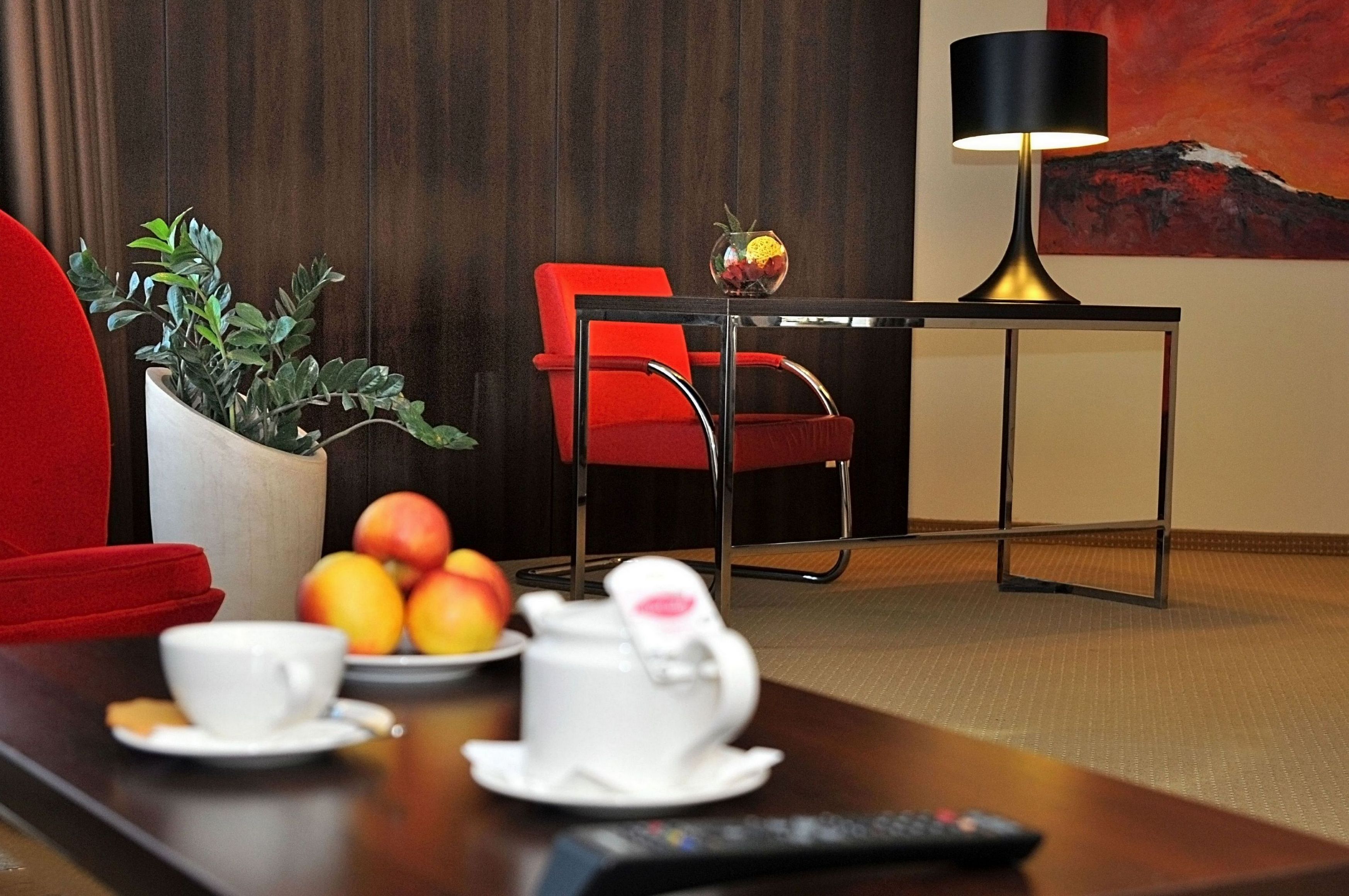 A hotel room with a red chair, a table with a white teapot, a cup, and a bowl of fruit.