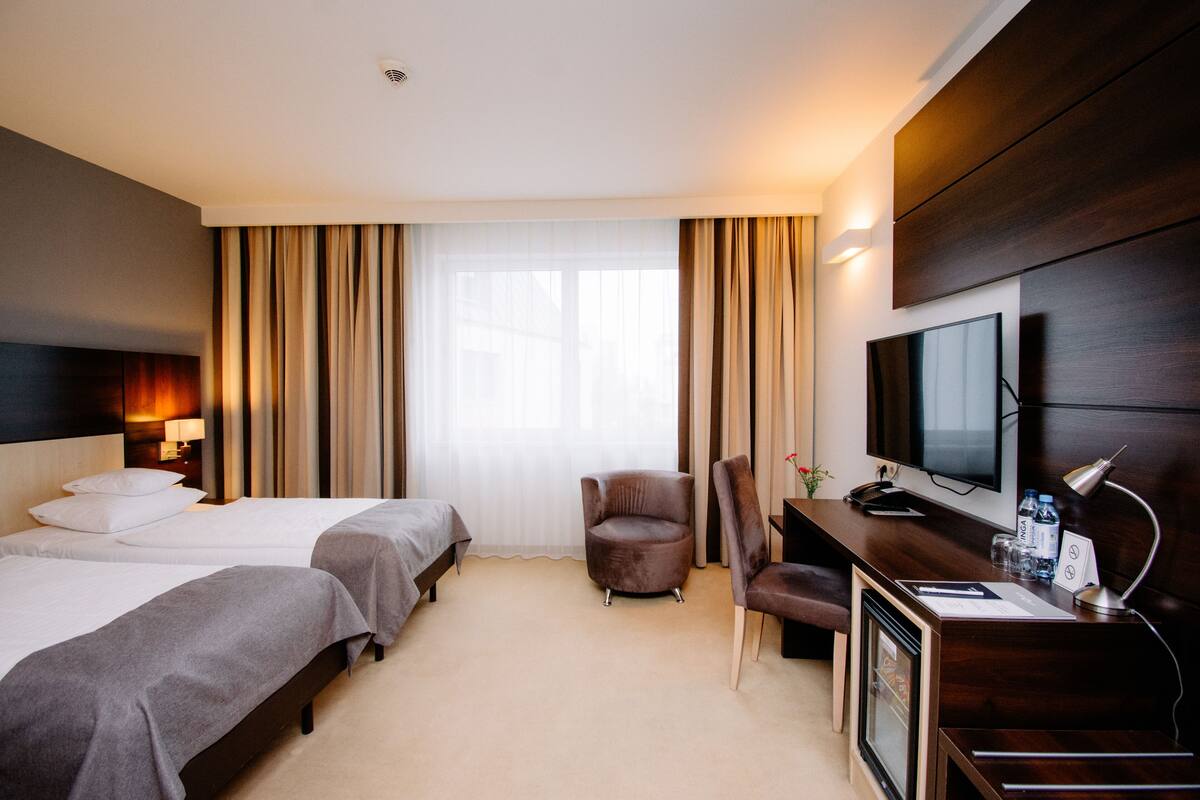Quarto de hotel com duas camas, uma escrivaninha com televisão, uma cadeira e uma janela grande com cortinas.