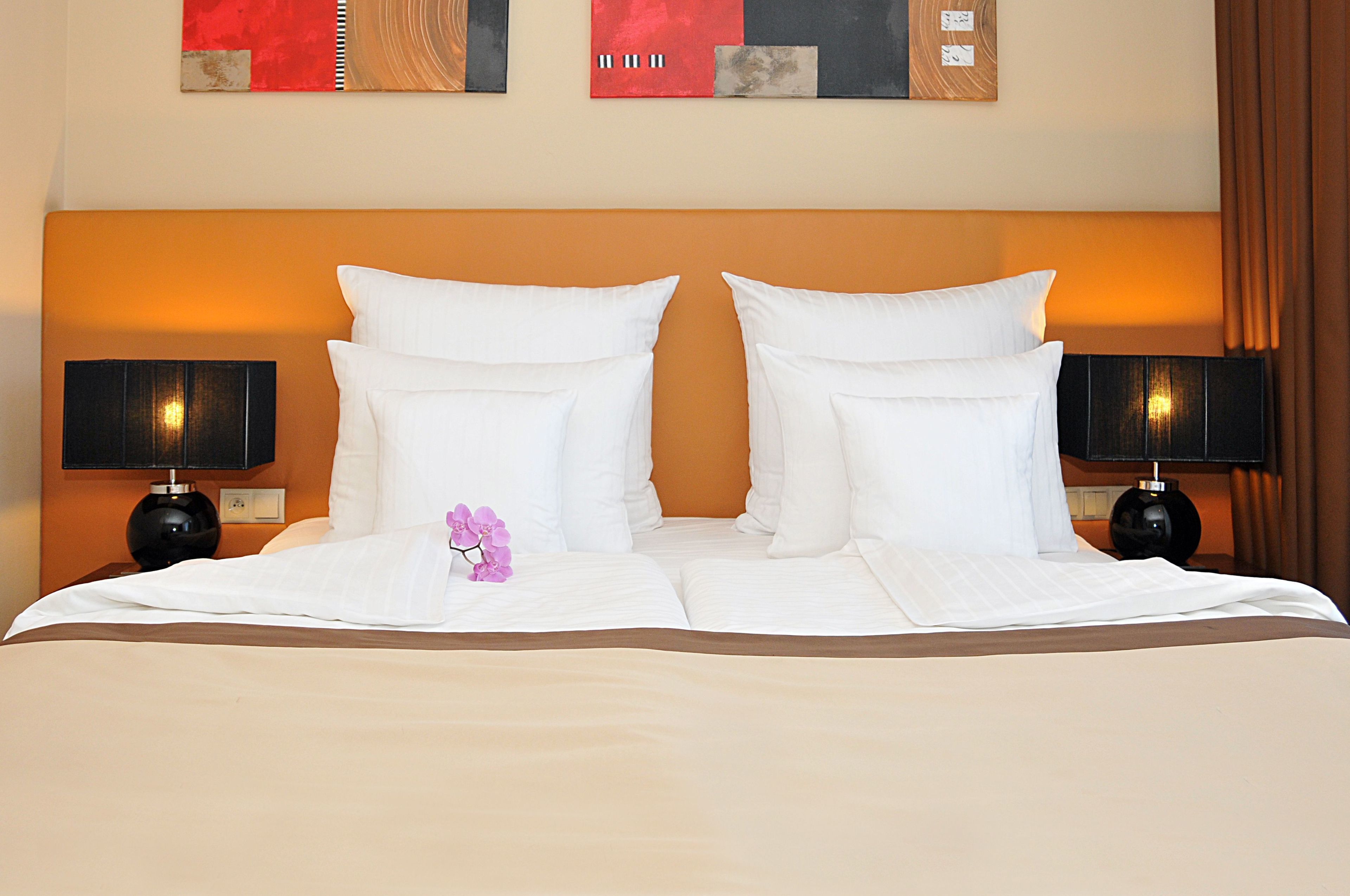 Una cama bien hecha con sábanas blancas y dos almohadas, flanqueada por dos mesitas de noche negras con lámparas, en una habitación con cabecera naranja y una obra de arte colgada en la pared.