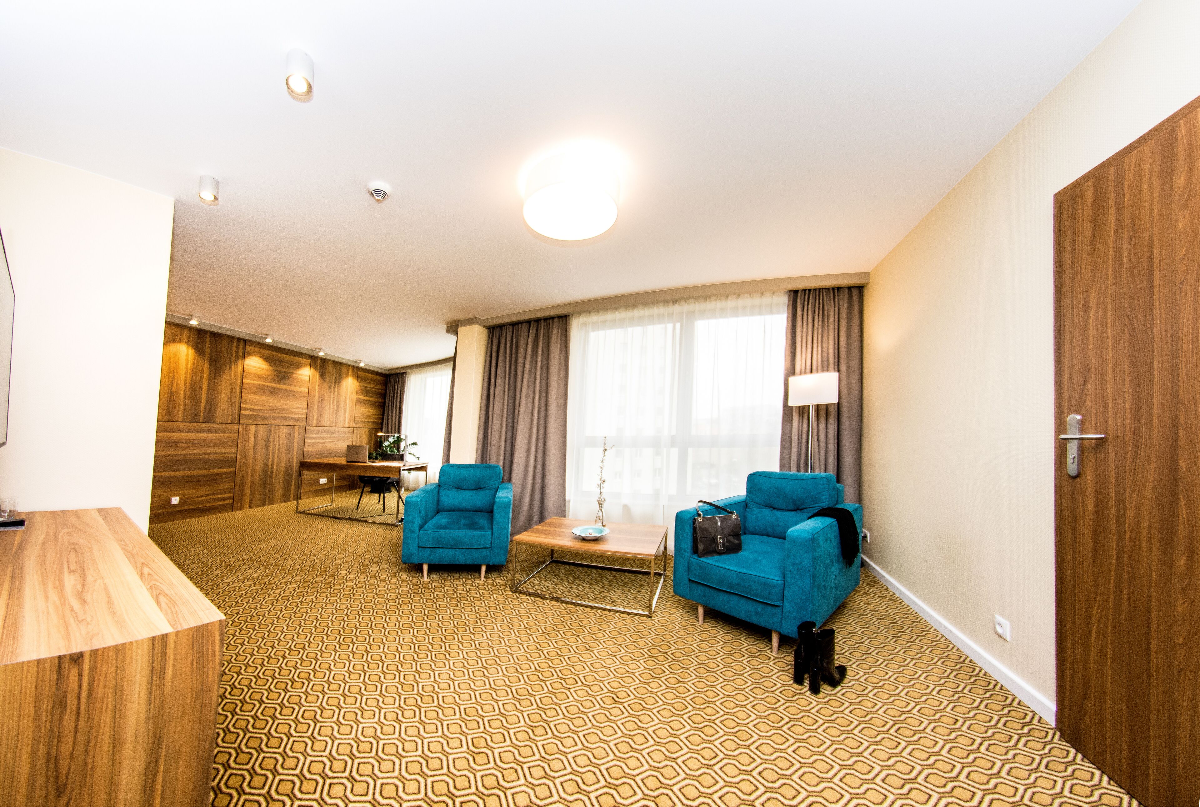 A hotel room with a patterned carpet, two blue armchairs, a wooden coffee table, and a large window with curtains.