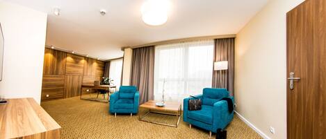 A hotel room with a patterned carpet, two blue armchairs, a wooden coffee table, and a large window with curtains.