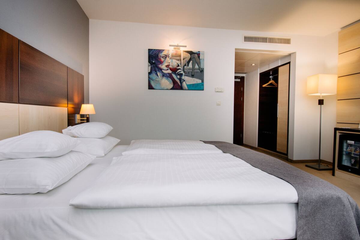 Quarto de hotel com uma cama grande, um quadro na parede, um guarda-roupa e uma mesa de cabeceira com abajur.