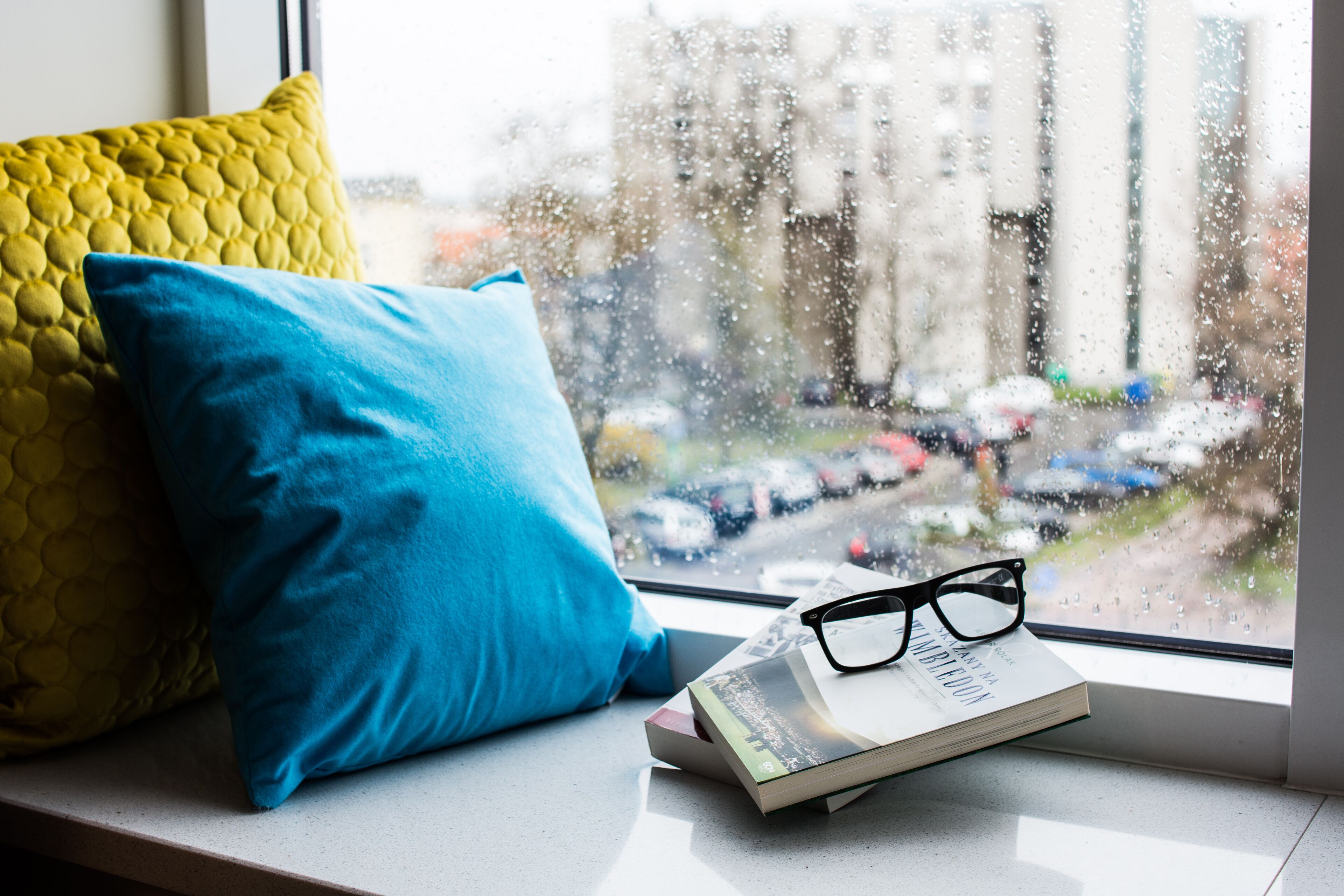 A window seat with a book and glasses, a yellow and blue pillow, and a view of parked cars and buildings outside.