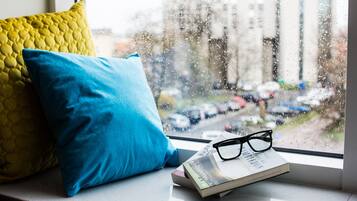 A window seat with a book and glasses, a yellow and blue pillow, and a view of parked cars and buildings outside.
