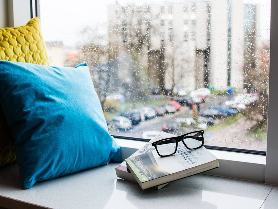A window seat with a book and glasses, a yellow and blue pillow, and a view of parked cars and buildings outside.