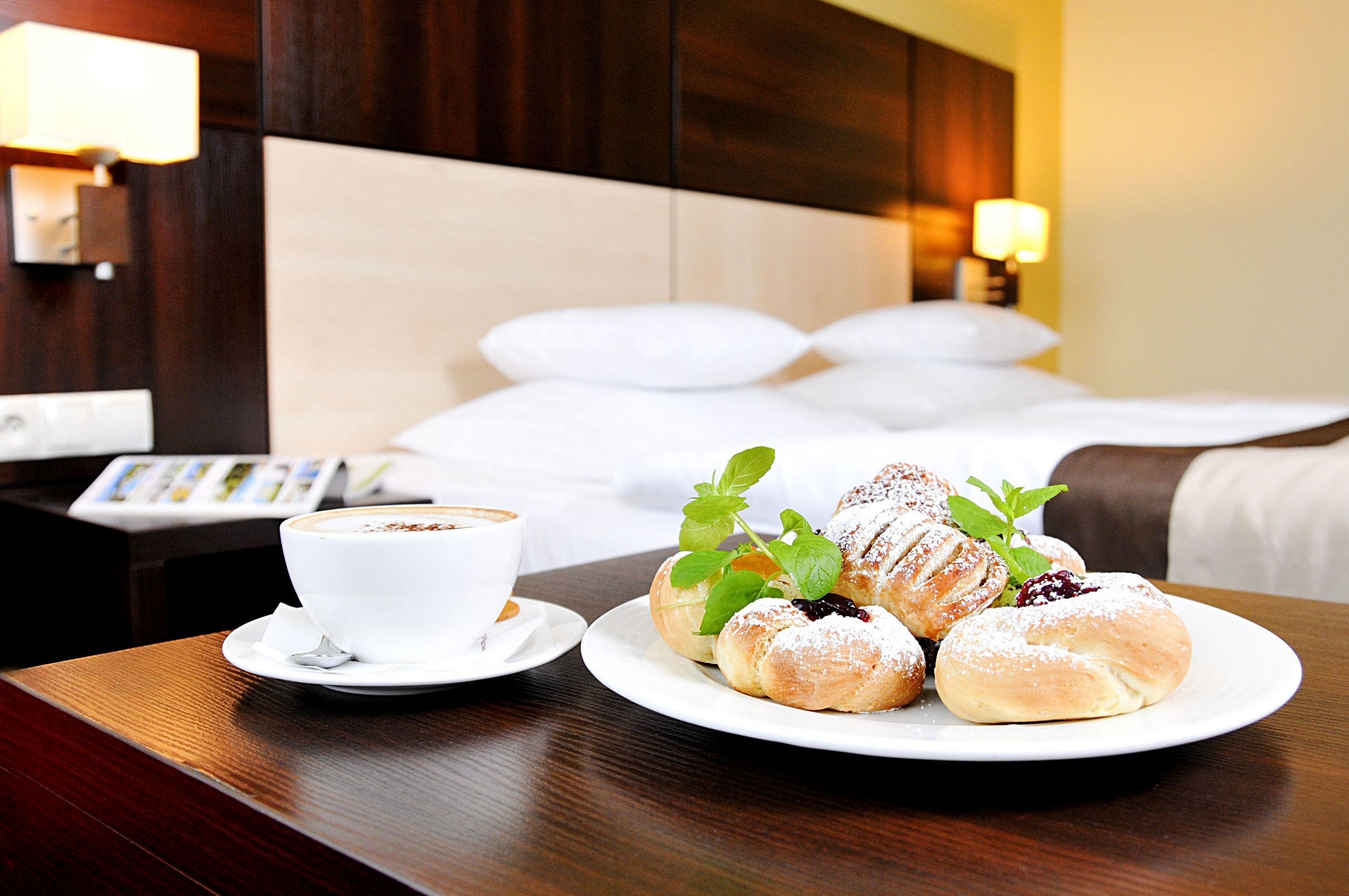 Habitación de hotel con una cama bien hecha, una taza de café sobre una mesa y un plato de pasteles.
