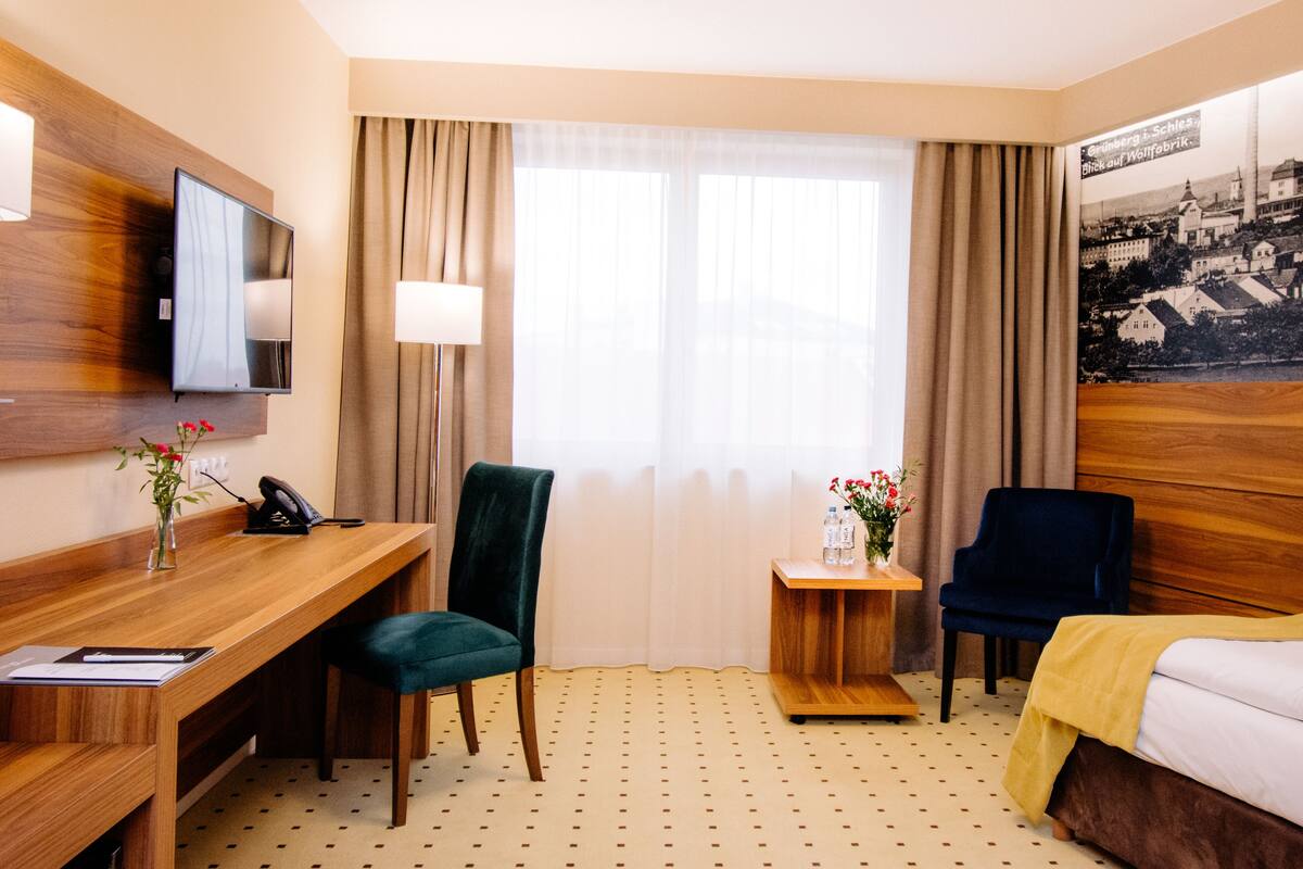 Quarto de hotel com escrivaninha de madeira, uma cadeira, uma cama, uma mesinha de cabeceira com abajur e uma televisão na parede.