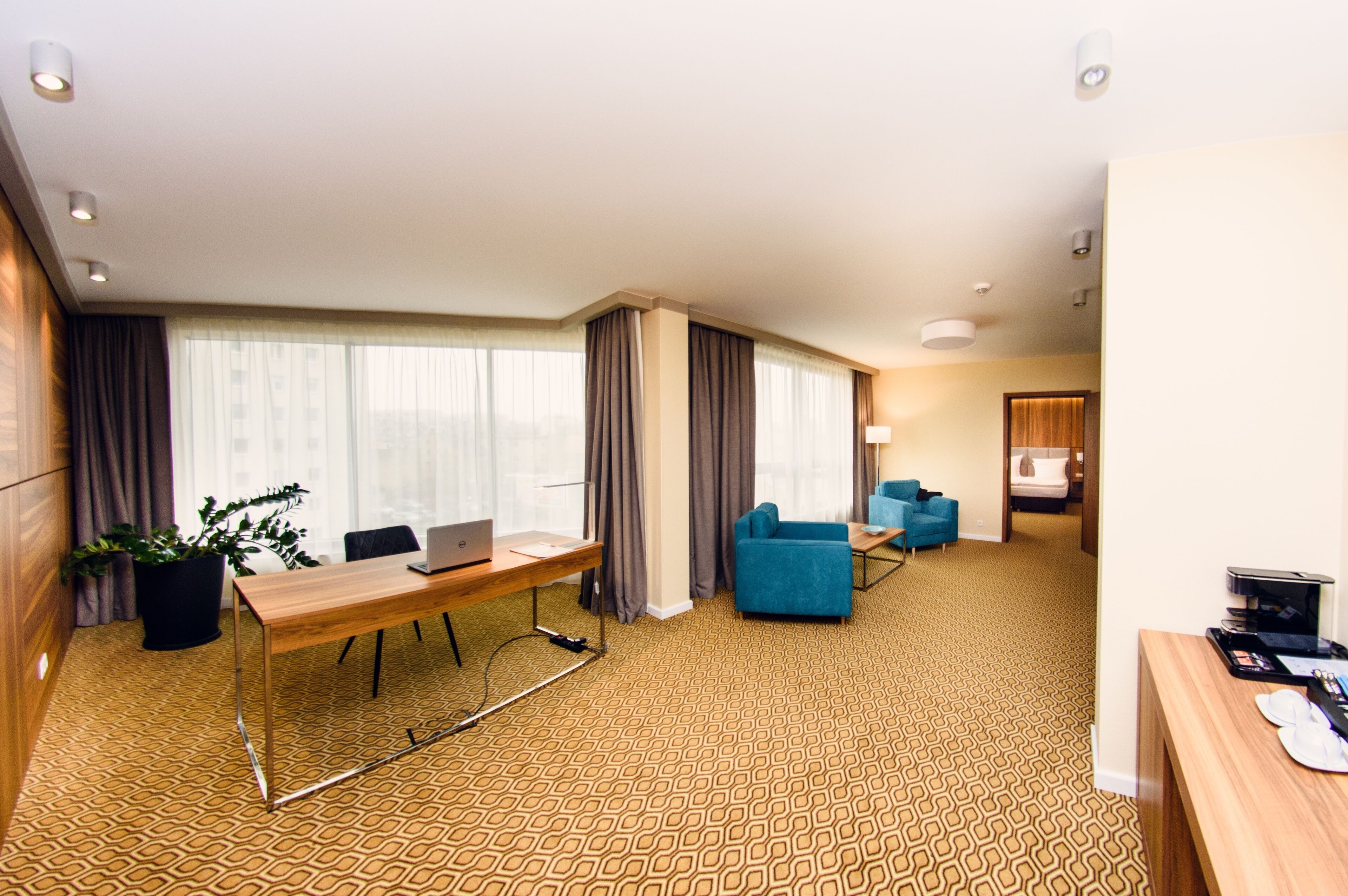 A hotel room with a wooden desk, two blue armchairs, a large window with curtains, and a patterned carpet.