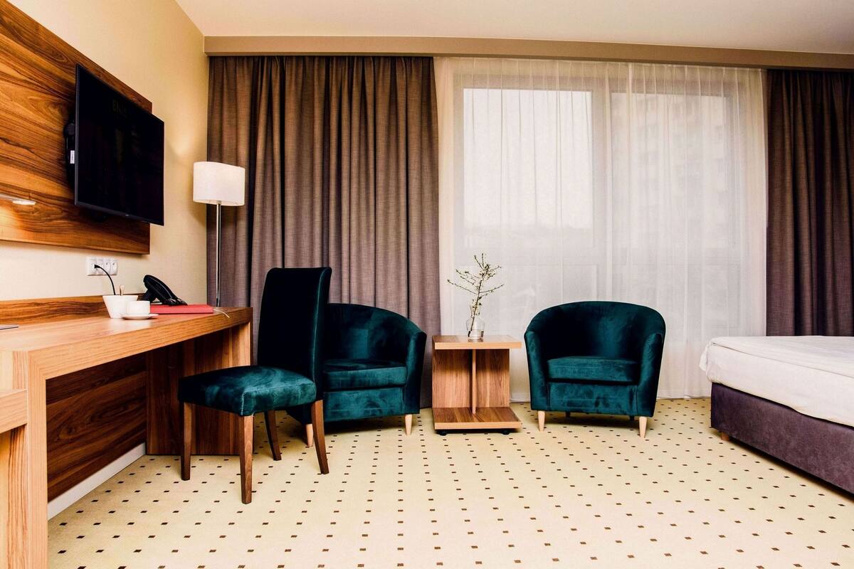 Quarto de hotel com uma escrivaninha de madeira, duas poltronas verde-escuras, uma mesinha de apoio, uma cama e uma televisão fixada na parede.