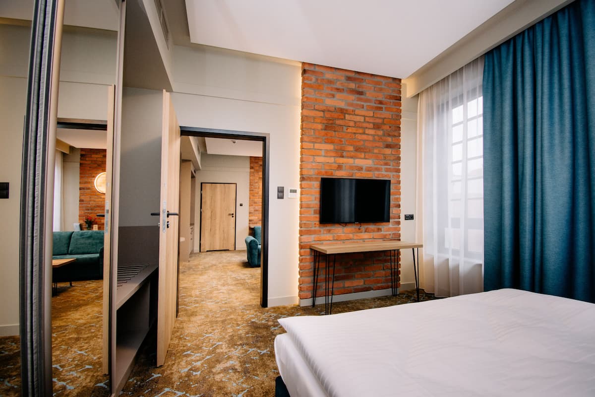 Quarto de hotel com parede de tijolos decorativa, televisão de tela plana, cama, escrivaninha e vista para um sofá verde e uma porta de madeira.