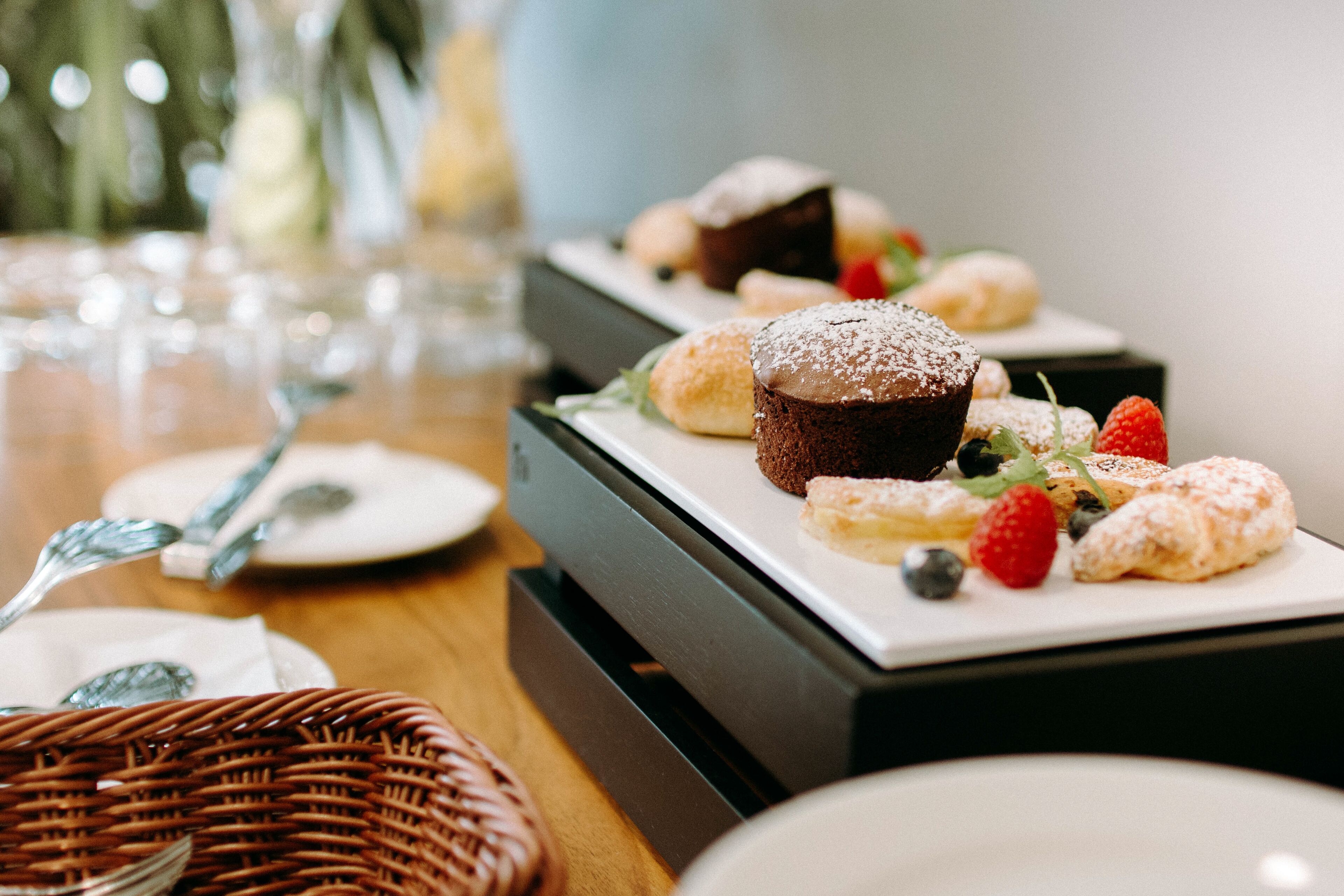 Une table dressée avec des assiettes, un panier et une variété de pâtisseries, notamment un muffin au chocolat, des croissants et un dessert aux fraises et aux myrtilles.