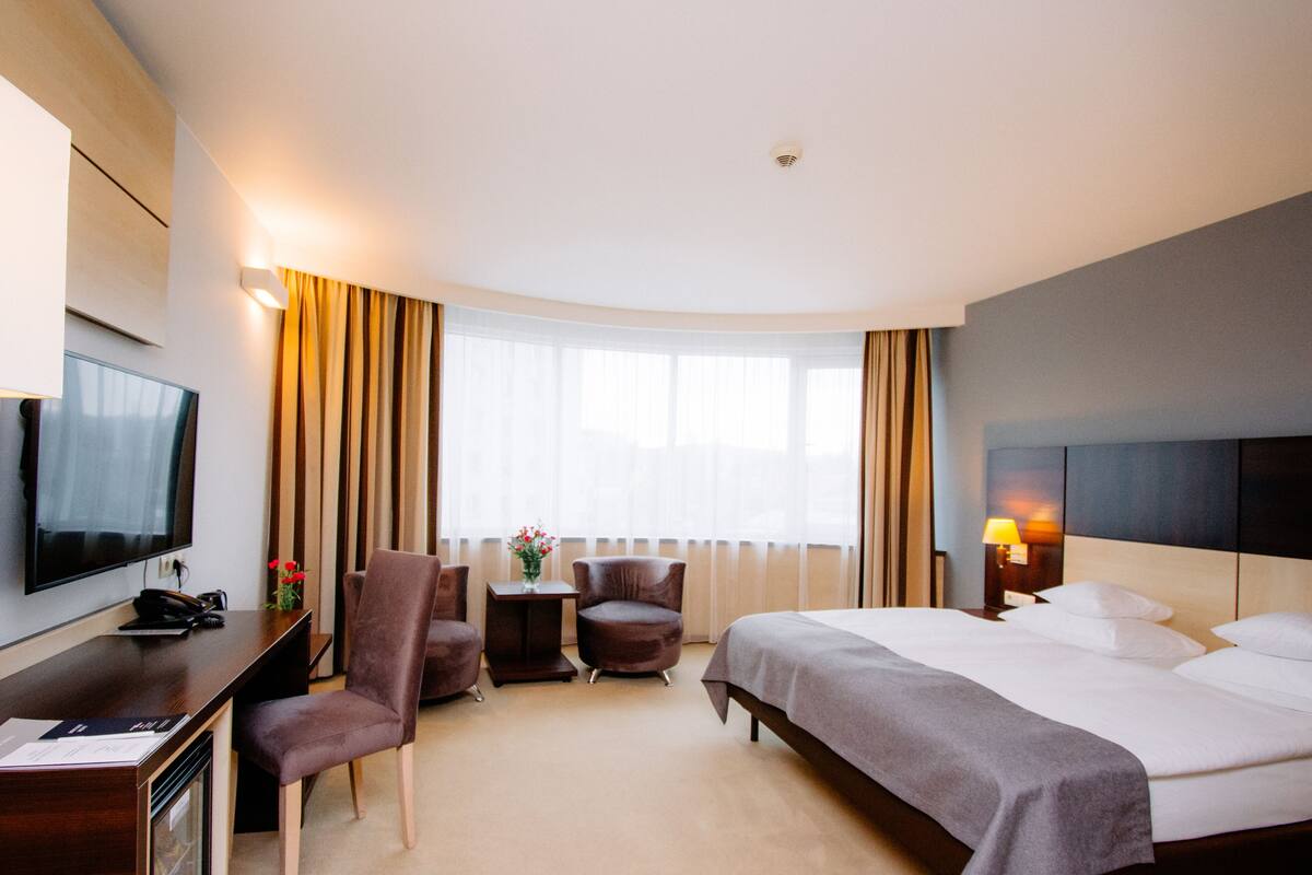 Quarto de hotel com cama, mesa de escritório, computador, televisão e duas cadeiras.
