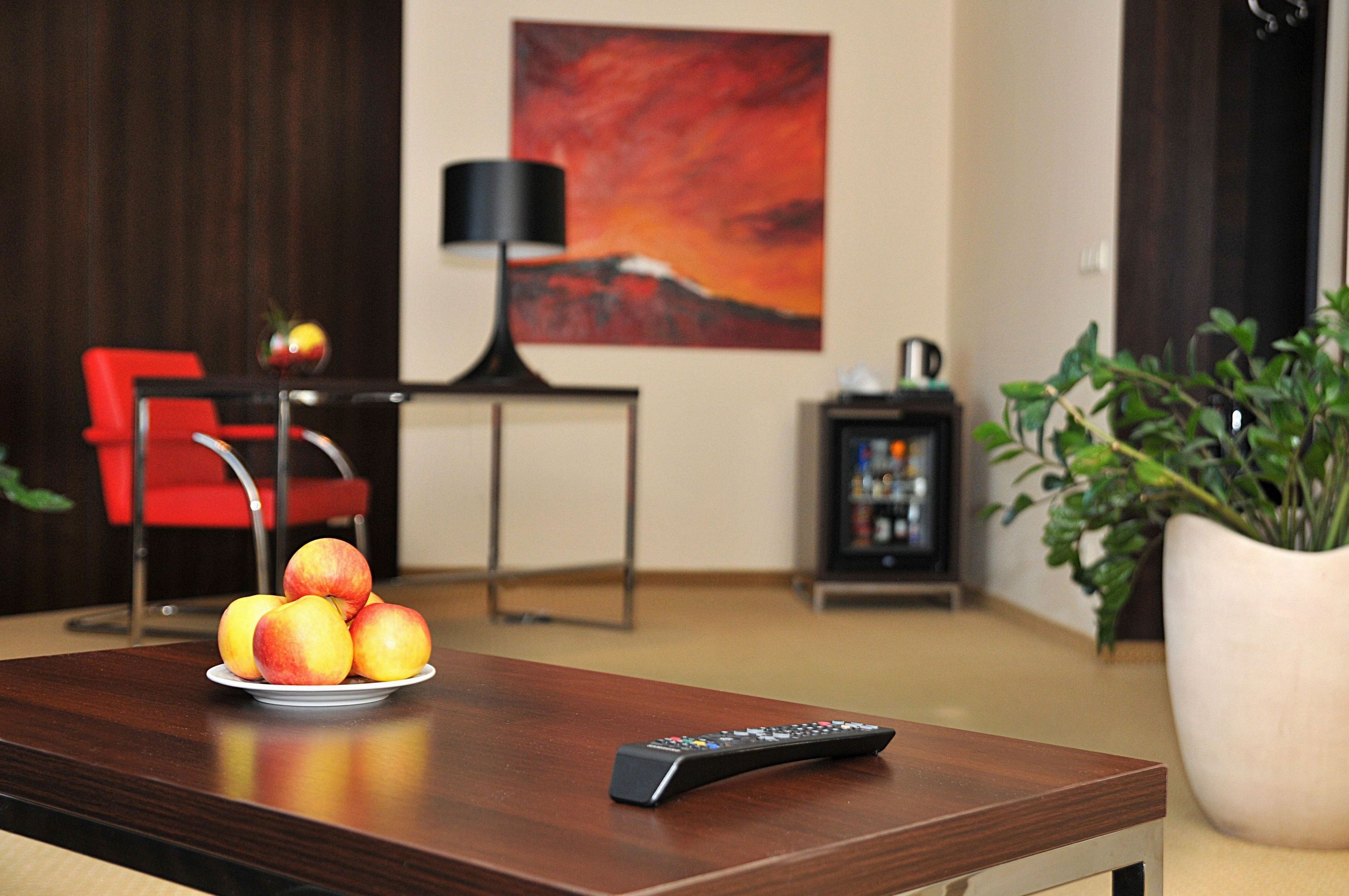 A wooden table with a remote control and a plate of fruit. A red chair and a black lamp are in the background.