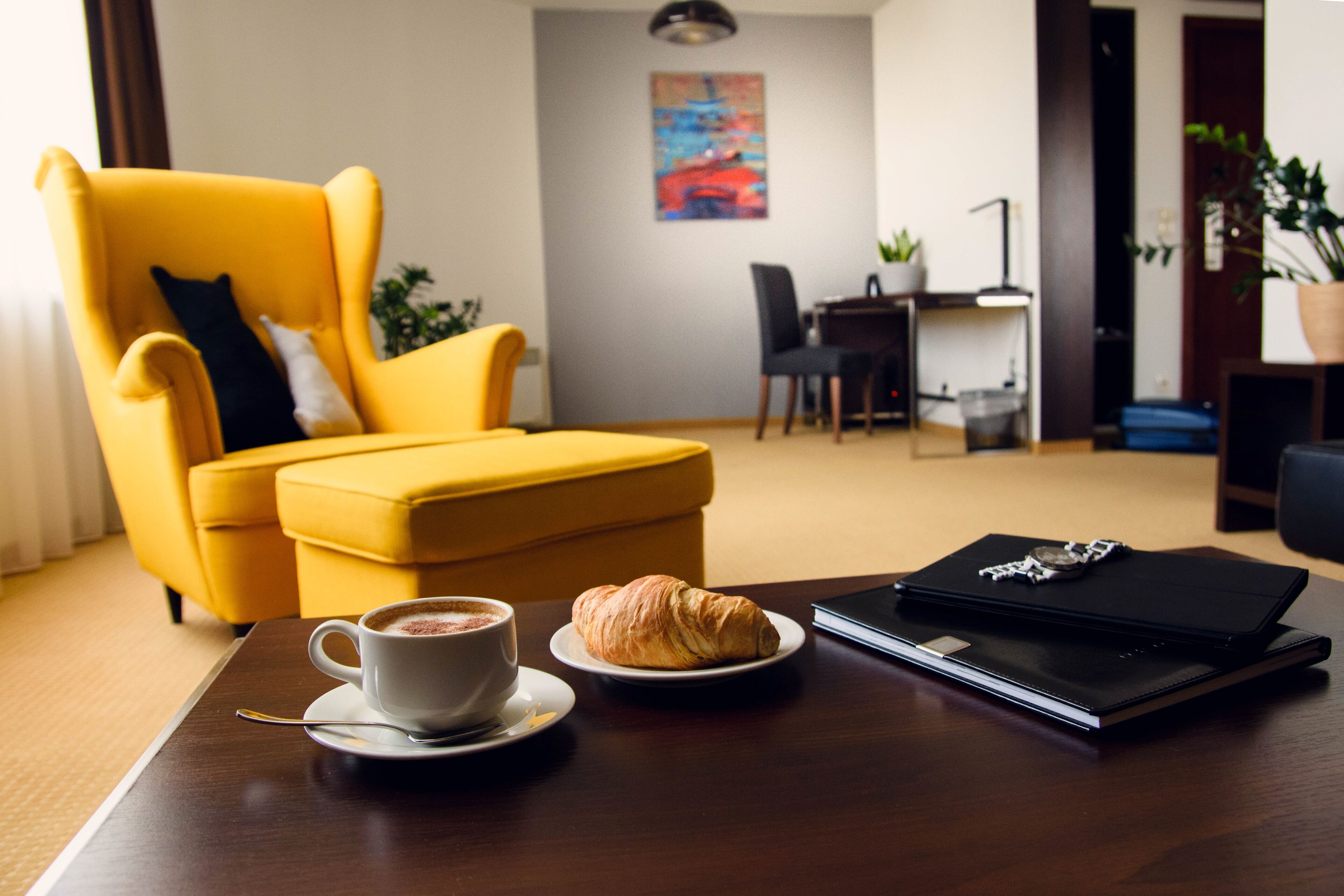 Ett hotellrum med en gul fåtölj, ett matbord med en croissant och en kopp kaffe, och en bärbar dator på bordet.