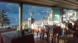 A restaurant with outdoor seating overlooking a lake and mountains. Tables are set with white tablecloths and metal chairs. - San Marino (Laglio)