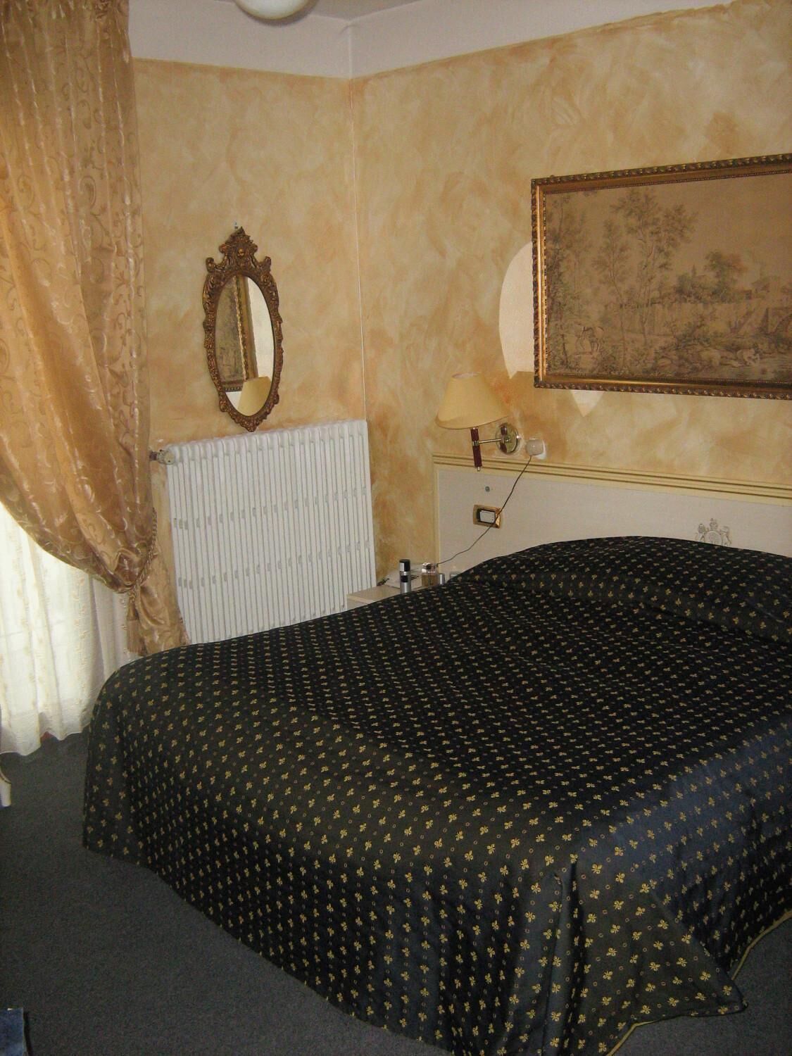A hotel room with a bed, a radiator, a mirror, a lamp, and a painting on the wall.