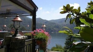 A terrace with a view of a lake and mountains, featuring a wooden railing, potted plants, and hanging lanterns. - San Marino (Laglio)