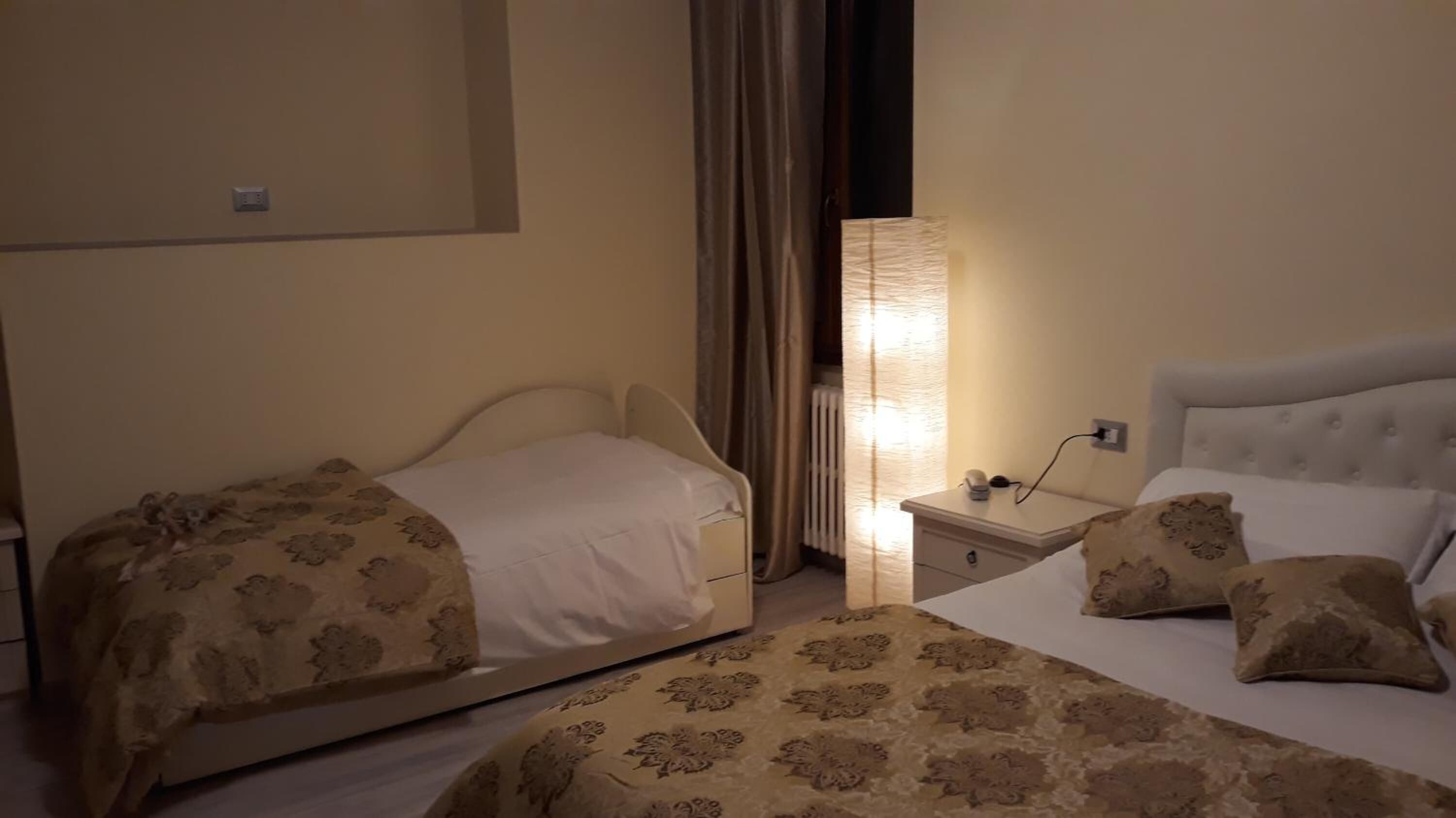 A hotel room with two beds, a bedside table with a lamp, and a radiator.