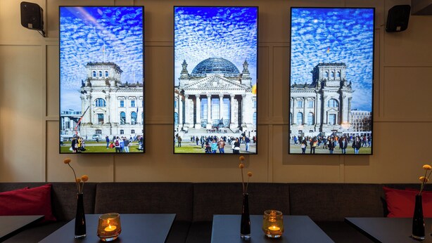 A restaurant with three large wall-mounted TVs displaying buildings, a dark-colored interior, and candles on the tables. - Leonardo Royal Hotel Berlin Alexanderplatz (Berlin)